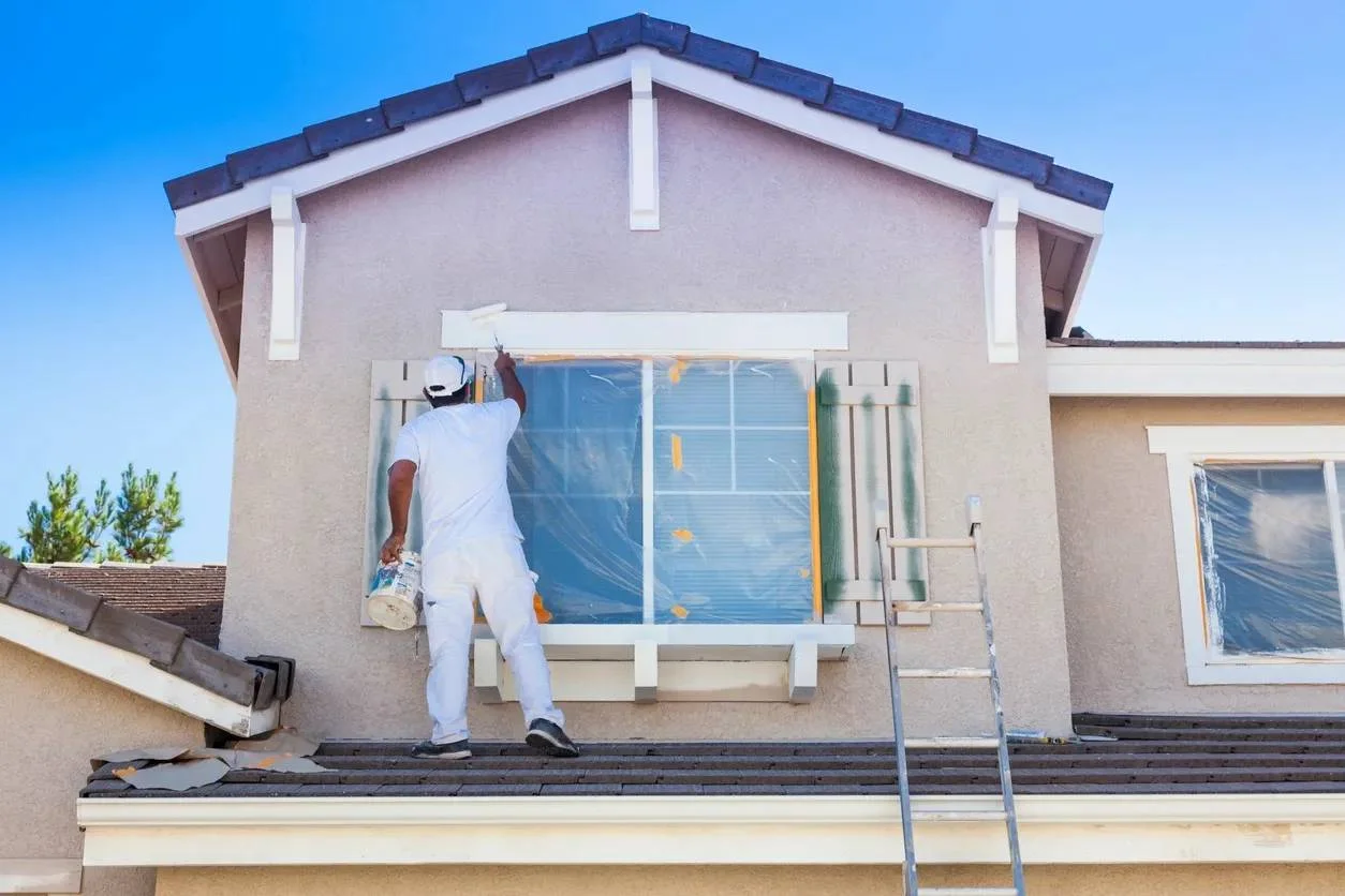 Painter in white uniform applying exterior paint to stucco house facade with white trim