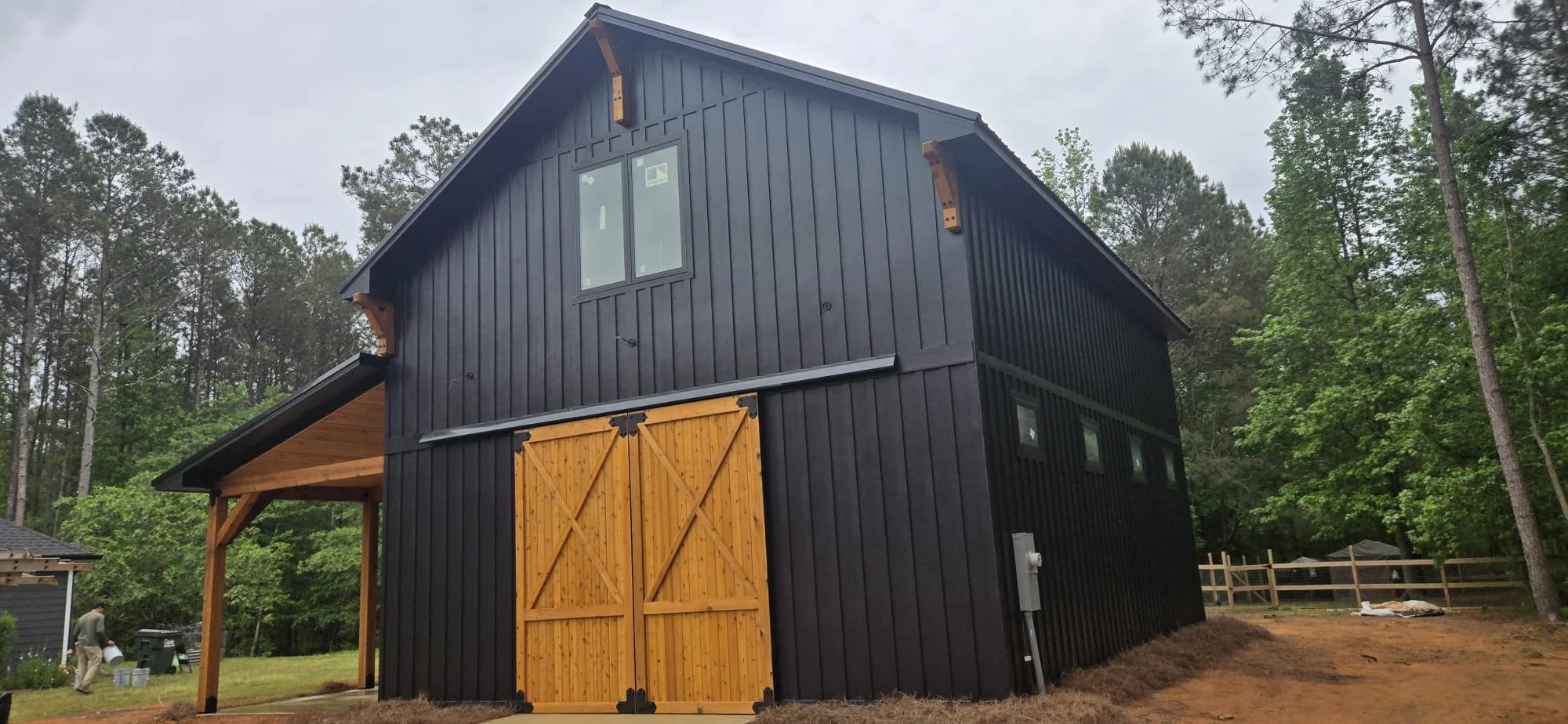 Black metal barn with wooden doors and gable windows surrounded by pine trees